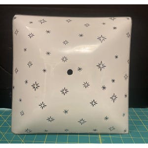 Puede incluir: Pantalla de lámpara cuadrada, blanquecina, con un patrón de estrellas negras. La pantalla tiene un agujero central y probablemente sea para una lámpara de techo. Las estrellas son de varios tamaños y estilos.