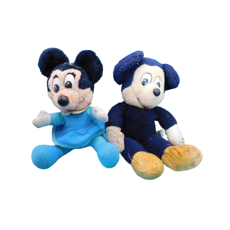 K&ouml;nnte beinhalten: Zwei Vintage-Mickey-Mouse-Pl&uuml;schtiere. Eines tr&auml;gt ein hellblaues Outfit, das andere ein dunkelblaues Outfit. Beide haben schwarze Ohren, wei&szlig;e Gesichter und braune F&uuml;&szlig;e. Die Spielzeuge sitzen auf wei&szlig;em Hintergrund.