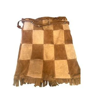 May include: A rectangular suede bag featuring a checkerboard pattern of alternating dark brown and light tan squares. The bag includes a thin brown leather strap with metal rings at the top and a fringe trim along the bottom edge of the piece.