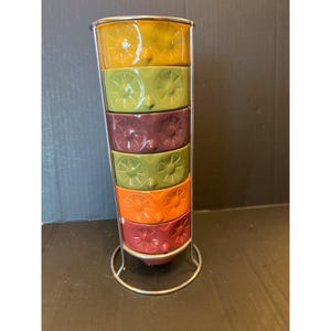 以下が含まれることがあります： 金属製のスタンドに入った、6つのカラフルなセラミックボウルのセット。ボウルは垂直に積み重ねられており、それぞれ異なる色（黄色、緑、紫、緑、オレンジ、赤）です。各ボウルには花の模様があります。金属製のスタンドはシルバーです。