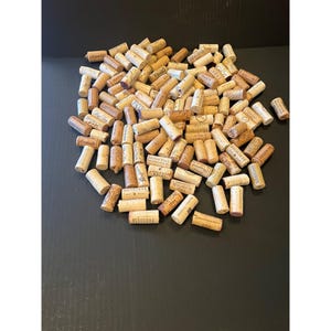 May include: A large pile of used wine corks, varying in size and shape, with visible text and markings. The corks are primarily light brown with some darker variations, suggesting different types of wine.
