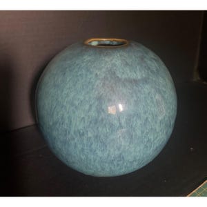 May include: A round, teal-colored vase with a speckled texture and a gold-colored rim. The vase has a smooth, glossy finish and a spherical shape. This decorative vase is suitable for displaying flowers or as a standalone art piece.