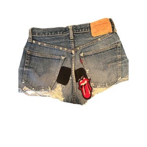 May include: Distressed denim shorts with a frayed hem. Features a black patch and a red and white Rolling Stones tongue patch. The shorts have a Levi Strauss & Co. label.