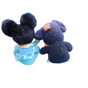 K&ouml;nnte beinhalten: Zwei Vintage-Pl&uuml;schtiere, eine Maus mit gro&szlig;en schwarzen Ohren und einem blauen Kleid und ein dunkelblauer B&auml;r mit einem Flicken auf dem R&uuml;cken. Die Spielzeuge sind dem Betrachter abgewandt, wobei die Maus die Hand des B&auml;ren h&auml;lt.