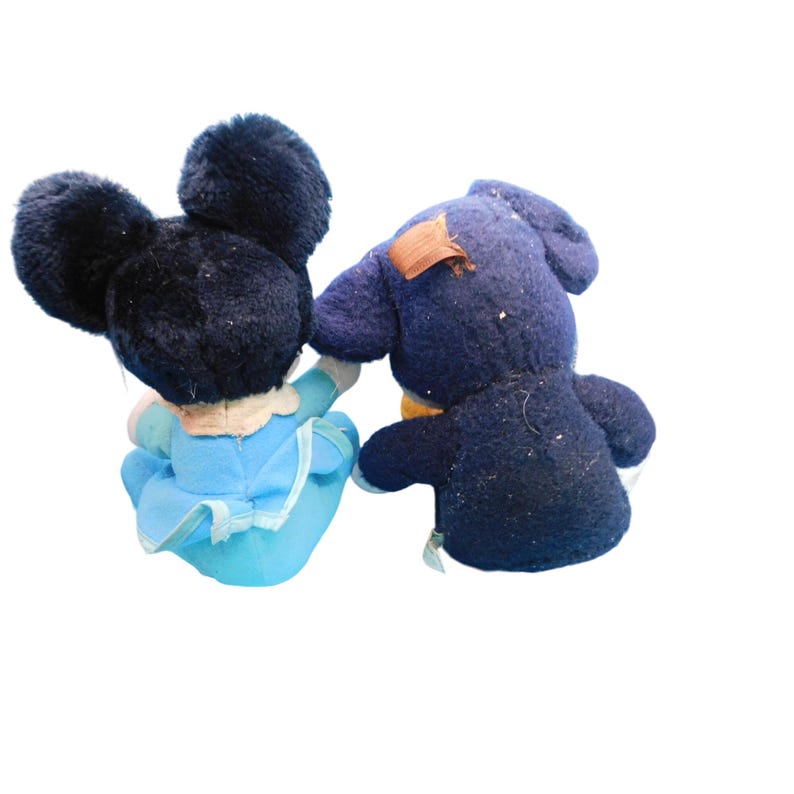 K&ouml;nnte beinhalten: Zwei Vintage-Pl&uuml;schtiere, eine Maus mit gro&szlig;en schwarzen Ohren und einem blauen Kleid und ein dunkelblauer B&auml;r mit einem Flicken auf dem R&uuml;cken. Die Spielzeuge sind dem Betrachter abgewandt, wobei die Maus die Hand des B&auml;ren h&auml;lt.