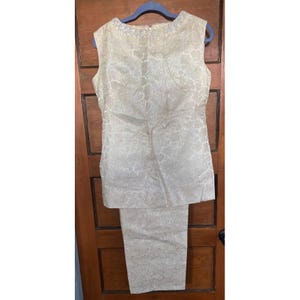 May include: A vintage two-piece outfit in a light cream color. The sleeveless top and matching pants are made of a textured fabric with a floral pattern. The top has a high neckline and a tailored fit.