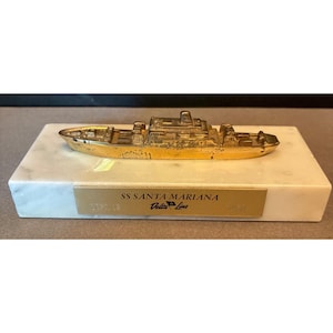 May include: A gold-colored metal ship model, SS Santa Mariana, mounted on a rectangular marble base. The base has a gold plaque with the ship's name and the words "Delta Line". The model is a detailed representation of a ship.