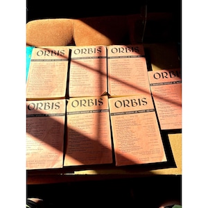 May include: A collection of vintage "ORBIS" journals, a quarterly journal of world affairs, with a salmon-colored cover. The journals are arranged on a dark surface, with sunlight casting shadows across them. The text on the cover is in a serif font.