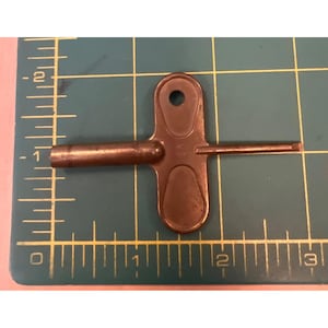 May include: A copper-colored metal radiator key with a T-shaped handle and a cylindrical shaft, positioned against a green cutting mat with a visible ruler showing a length of approximately 2.5 inches from tip to handle edge.