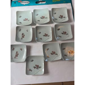May include: Set of ten small, square, light blue ceramic dishes with a pine cone design. The dishes are arranged in two rows of five. The dishes are labeled "Stickies".