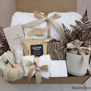May include: An autumn-themed gift box filled with self-care items. Includes a white cable-knit blanket, Shea-Socks, Gel-Ohh! Milk & Honey spa bath, a mug, and a decorative pumpkin. The box is tied with a gold ribbon and includes the text "Autumn Serenity Box".