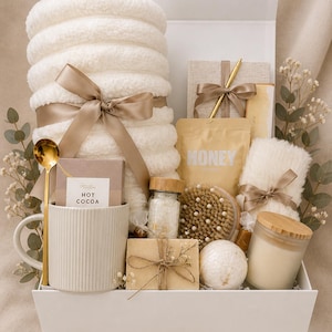 May include: A white gift box containing a rolled white plush blanket tied with a beige ribbon, a ribbed mug with a gold spoon, a hot cocoa packet, a honey pouch, a notebook with a gold pen, a bath brush, a candle, a soap bar, and a bath bomb.