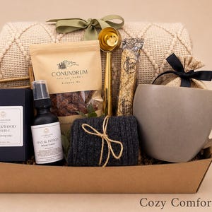May include: A gift basket containing a beige textured throw blanket with a green ribbon, a bag of Conundrum coffee, a gold coffee scoop, a Blackwood Reserve soap box, an Oat & Honey repair cream bottle, a dark grey knit item, and a grey mug.