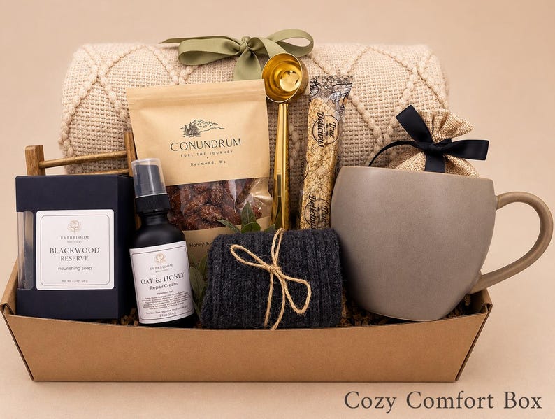 May include: A gift basket containing a beige textured throw blanket with a green ribbon, a bag of Conundrum coffee, a gold coffee scoop, a Blackwood Reserve soap box, an Oat & Honey repair cream bottle, a dark grey knit item, and a grey mug.