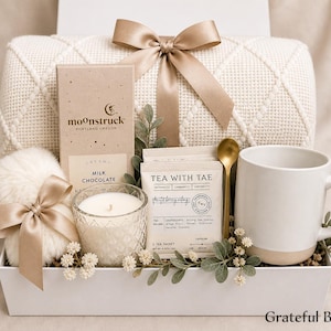 May include: A gift box containing a cream-coloured knit blanket with a satin ribbon, a Moonstruck milk chocolate bar, a white candle in a textured glass, Tea with Tae tea sachets, a gold spoon, a white mug, and a faux fur pom-pom with a ribbon.