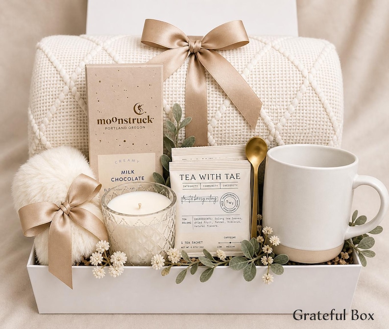 May include: A gift box containing a cream-coloured knit blanket with a satin ribbon, a Moonstruck milk chocolate bar, a white candle in a textured glass, Tea with Tae tea sachets, a gold spoon, a white mug, and a faux fur pom-pom with a ribbon.