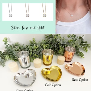 K&ouml;nnte beinhalten: Drei Halsketten in Silber, Ros&eacute;gold und Gold mit einem kleinen runden Anh&auml;nger. Unter den Halsketten befinden sich drei herzf&ouml;rmige Tabletts in Silber, Gold und Ros&eacute;gold. Jedes Tablett hat einen Teelichthalter und eine Kerze auf einer wei&szlig;en Oberfl&auml;che. Der Text "Kerzen haben einen Vanilleduft" befindet sich unter den Tabletts.