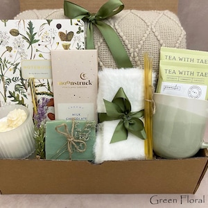 May include: A gift box containing a floral notebook, a Moonstruck milk chocolate bar, a white faux fur item with a green ribbon, two Tea With Tae tea bags, a sage green ceramic mug, a ribbed glass candle, a honey dipper, and a beige knit blanket.
