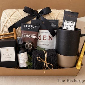 May include: A gift basket with a black bow, containing a black and beige mug, a bag of coffee, a gold spoon, a bar of soap, a bottle of spray, a bag of almonds, a face mask, and a small blanket. The Recharge Box is written on the bottom.
