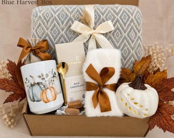 May include: A Blue Harvest Box gift set containing a blue and white diamond-patterned blanket with a cream ribbon, a white mug with pumpkin illustrations, a gold spoon, a milk chocolate bar, a white faux fur item, and a white ceramic pumpkin.