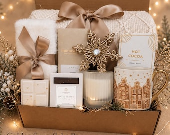 May include: A gift box containing a white faux fur item with a brown ribbon, a brown bar, a gold snowflake ornament, a white Hot Cocoa packet, a white Oat & Honey soap bar, a white wax melt, a ribbed glass candle, and a mug with gold house designs.