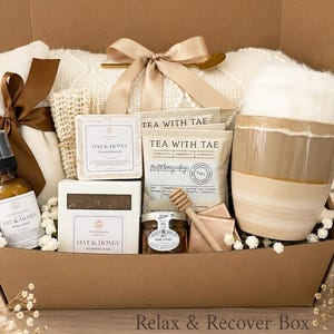 May include: A gift box containing a ceramic mug with a beige and white glaze, two Tea With Tae tea bags, a small jar of honey with a wooden dipper, two Oat & Honey soap bars, a spray bottle of body lotion, and a white textured blanket with a bow.