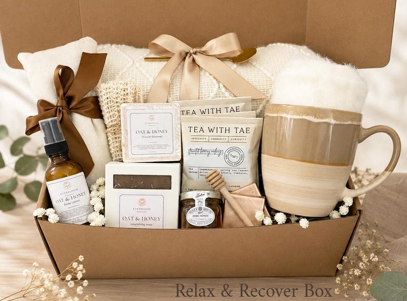 May include: A gift box containing a ceramic mug with a beige and white glaze, two Tea With Tae tea bags, a small jar of honey with a wooden dipper, two Oat & Honey soap bars, a spray bottle of body lotion, and a white textured blanket with a bow.