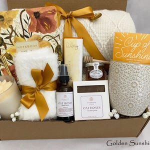 May include: A gift box containing self-care products. It features a notebook, a candle, a mug with a "Cup of Sunshine" tea packet, Oat Honey body lotion, Oat Honey soap, a jar of honey, and a soft blanket. The box is tied with a gold ribbon.