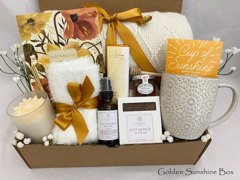 May include: A gift box containing self-care products. It features a notebook, a candle, a mug with a "Cup of Sunshine" tea packet, Oat Honey body lotion, Oat Honey soap, a jar of honey, and a soft blanket. The box is tied with a gold ribbon.