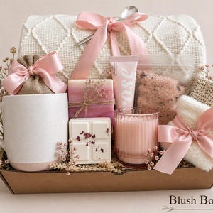 May include: A gift set in a cardboard box containing a white ceramic mug, a cream-colored knit blanket tied with a pink ribbon, a pink candle, two soap bars, a tube labeled rose, a pouch of bath salts, and a small white towel with a pink ribbon.