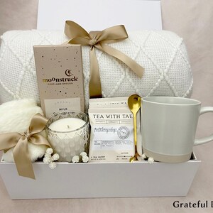 May include: A white gift box containing a cream-coloured blanket, a mug, a candle, a box of chocolate, tea packets, a gold spoon, and a fluffy item, all tied with beige ribbons. The box is labelled "Grateful Box".