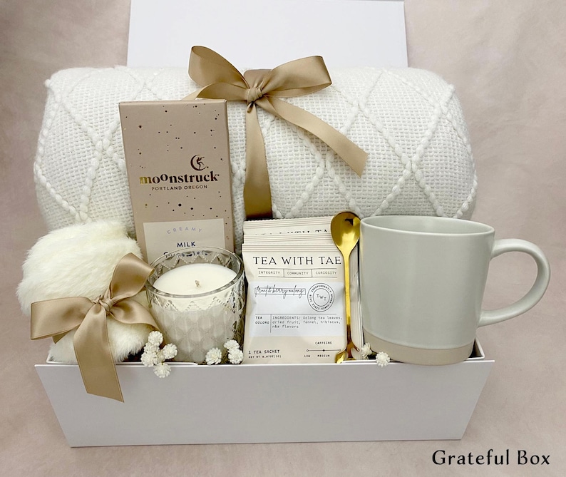 May include: A white gift box containing a cream-coloured blanket, a mug, a candle, a box of chocolate, tea packets, a gold spoon, and a fluffy item, all tied with beige ribbons. The box is labelled "Grateful Box".