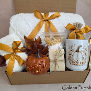 May include: A gift box containing a white knit blanket with a gold ribbon, a white mug with a pumpkin illustration, a gold spoon, a ceramic pumpkin lantern, a bar of soap, a small burlap bag with a gold ribbon, and artificial brown autumn leaves.