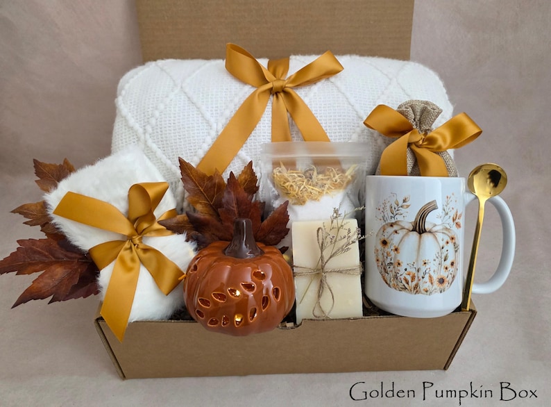 May include: A gift box containing a white knit blanket with a gold ribbon, a white mug with a pumpkin illustration, a gold spoon, a ceramic pumpkin lantern, a bar of soap, a small burlap bag with a gold ribbon, and artificial brown autumn leaves.