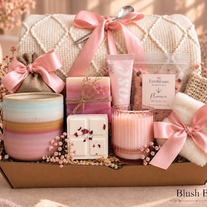 May include: A gift set in a cardboard box containing a ceramic mug with pink and tan bands, a pink ribbon-tied burlap pouch, a pink-toned soap bar, a rose-labeled tube, Everbloom Botanicals Plumeria bath salts, a wax melt, a pink candle, and a white fuzzy item.