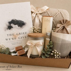 May include: A gift box filled with a book titled "The Grief We're Given", a candle, a mug, chocolate, and other items. The box is decorated with ribbons and sprigs of flowers. The overall color scheme is neutral with beige, brown, and white tones.