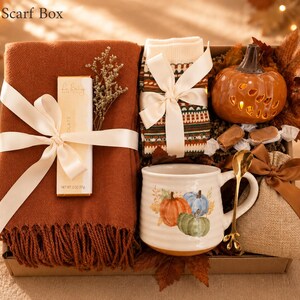 May include: A gift box labeled Cozy Scarf Box containing a rust-colored fringed scarf, a chocolate bar, patterned socks tied with a white ribbon, a ceramic pumpkin, a white mug with pumpkin illustrations, a gold spoon, and a small burlap bag.