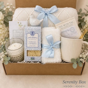 May include: A gift box containing a white textured blanket with a light blue ribbon, a white ceramic bird, a box of Unna Bakery vanilla cookies, a white candle in a patterned glass, a white mug, and a Tea With Tae packet with a wooden stirrer.