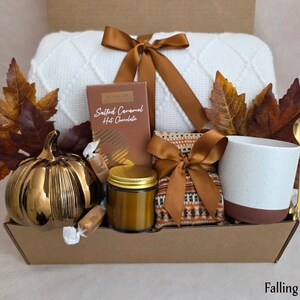May include: A gift box containing a white knit blanket tied with a brown ribbon, a box of Salted Caramel Hot Chocolate, a small jar with a gold lid, a patterned pouch, a white and brown mug, a gold spoon, and a metallic pumpkin, all with autumn leaves.