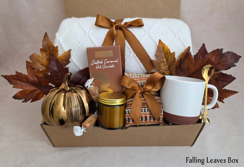 May include: A gift box containing a white knit blanket tied with a brown ribbon, a box of Salted Caramel Hot Chocolate, a small jar with a gold lid, a patterned pouch, a white and brown mug, a gold spoon, and a metallic pumpkin, all with autumn leaves.