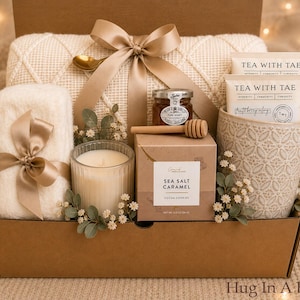 May include: A gift box containing a cream-colored textured blanket tied with a gold ribbon, a white candle in a ribbed glass, a jar of honey with a wooden dipper, a box of sea salt caramel cookies, and a mug with two Tea With Tae tea bags.