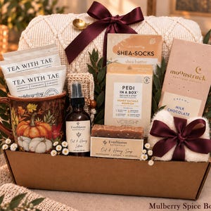May include: A gift basket containing a pumpkin-patterned mug, two Tea With Tae packets, a bottle of Everbloom Oat & Honey lotion, a bar of Everbloom soap, a Pedi in a Box kit, Shea-Socks, a Moonstruck milk chocolate bar, and a fuzzy white item with a dark ribbon.