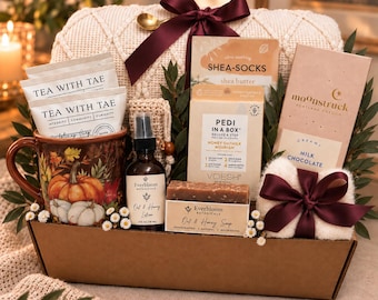 May include: A gift basket containing a pumpkin-patterned mug, two Tea With Tae packets, a bottle of Everbloom Oat & Honey lotion, a bar of Everbloom soap, a Pedi in a Box kit, Shea-Socks, a Moonstruck milk chocolate bar, and a fuzzy white item with a dark ribbon.