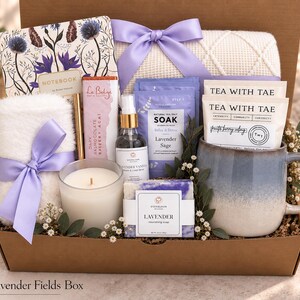 May include: A gift box containing a floral notebook, a white fuzzy towel with a lavender ribbon, a gold pen, a dark chocolate bar, a lavender vanilla spray, lavender sea salt soak, two tea bags, a white candle, and a lavender soap in a blue mug.