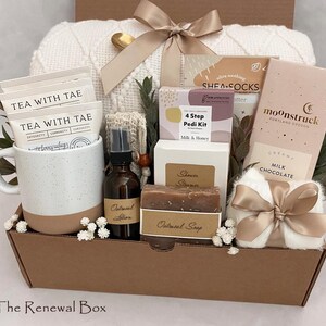 May include: A self-care gift basket containing a white speckled mug with tea bags, a brown lotion bottle, oatmeal soap, a chocolate bar, a white blanket, and other spa products. The basket is tied with a beige ribbon and presented in a brown box.
