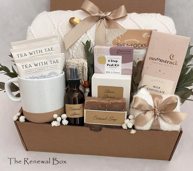 May include: A self-care gift basket containing a white speckled mug with tea bags, a brown lotion bottle, oatmeal soap, a chocolate bar, a white blanket, and other spa products. The basket is tied with a beige ribbon and presented in a brown box.