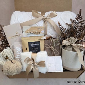 May include: A gift box containing a white cable knit blanket tied with a gold ribbon, a mug with a gold spoon, a small burlap bag, a white pumpkin, a bar of soap, a packet of Gel-Ohh! jelly spa bath, shea socks, a lotion tube, and dried fern leaves.