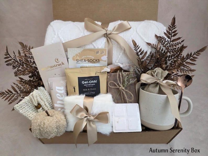 May include: A gift box containing a white cable knit blanket tied with a gold ribbon, a mug with a gold spoon, a small burlap bag, a white pumpkin, a bar of soap, a packet of Gel-Ohh! jelly spa bath, shea socks, a lotion tube, and dried fern leaves.