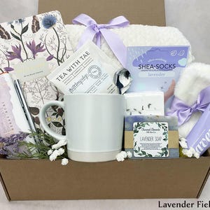 May include: A gift box containing lavender-scented products. It features a mug, tea, soap, shea socks, a journal, a pen, a chocolate bar, and a small towel. The box is tied with a purple ribbon and has the text "Lavender Fields Box".