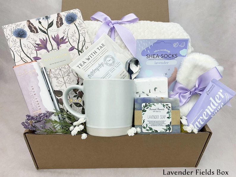 May include: A gift box containing lavender-scented products. It features a mug, tea, soap, shea socks, a journal, a pen, a chocolate bar, and a small towel. The box is tied with a purple ribbon and has the text "Lavender Fields Box".